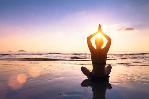 Person meditating on the beach at sunset with hands in prayer pose.