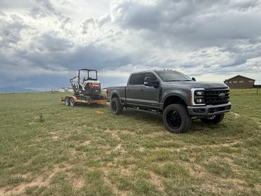 A gray pickup truck towing a trailer with a Bobcat excavator on a grassy field.