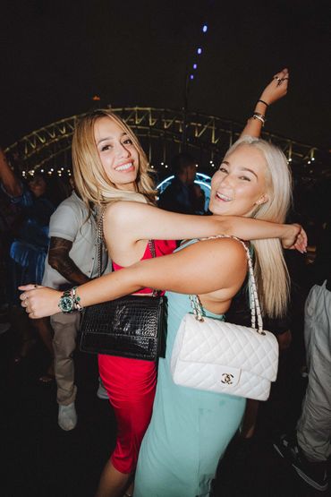 Two women joyfully hugging at a night event with a lit bridge in the background.