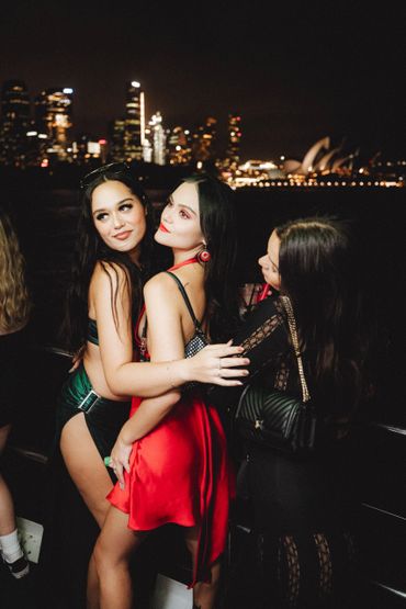 Three women posing and hugging outdoors at night with city lights in the background.