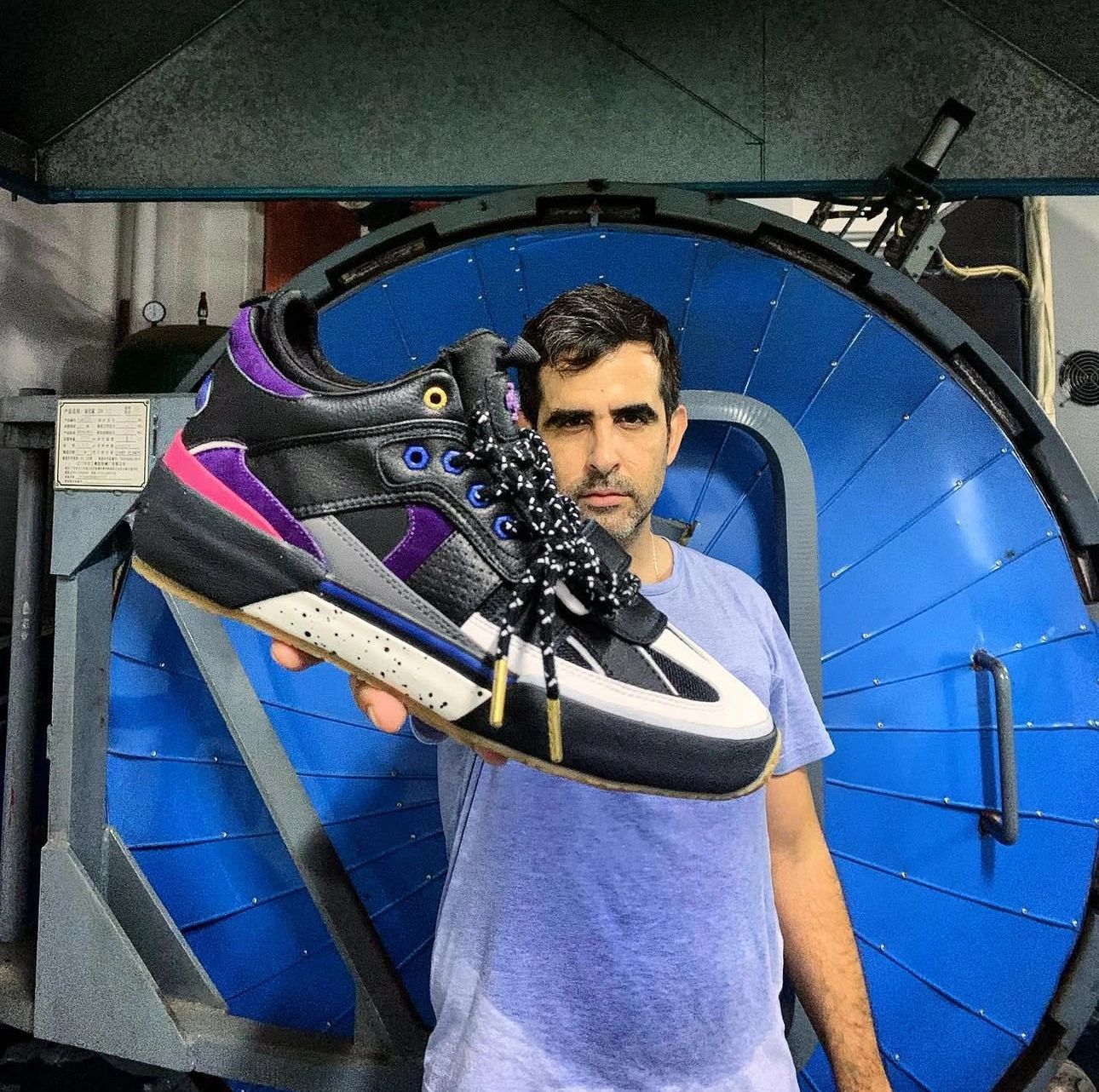Man holding a stylish black, purple, and white sneaker in front of a large blue industrial machine.