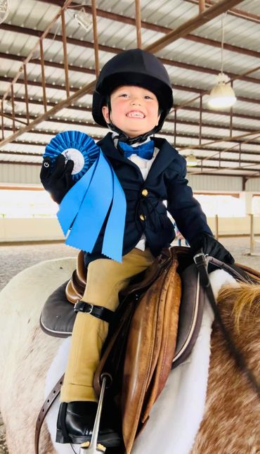 Little boy dressed in English riding attire holding a blue ribbon and wearing a big smile.