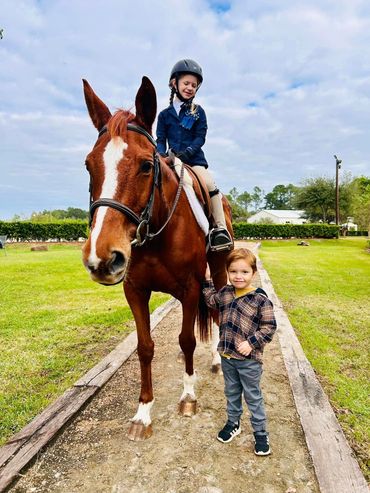 Little boy standing next to a chestnut horse with girl dressed in riding attire on top of horse.