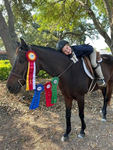Girl dressed in riding attire leaning down on horse's neck that is dressed with horse show ribbons.