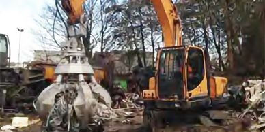 Excavator with claw attachment lifting scrap metal at a scrapyard.