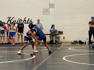 Two young wrestlers compete on a mat in a gymnasium with spectators and coaches watching.