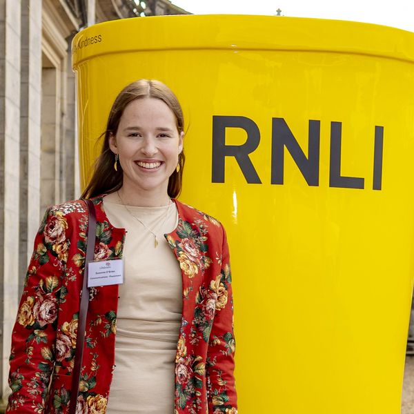 Woman in floral jacket smiles near large RNLI yellow bucket.