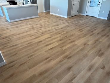 Empty room with light wood flooring and modern kitchen in the background.