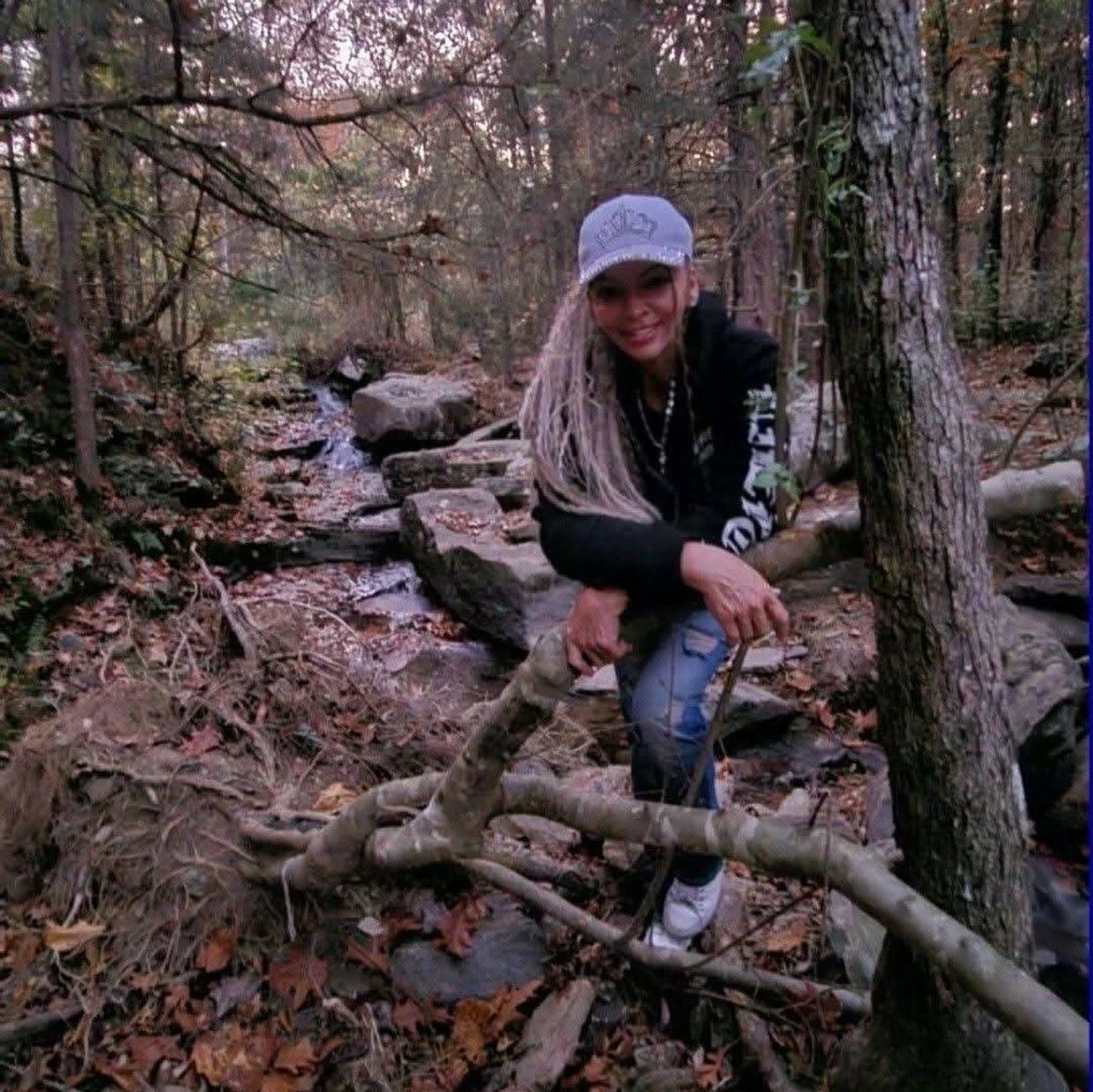 Woman in a cap smiling by a rocky stream in the woods.