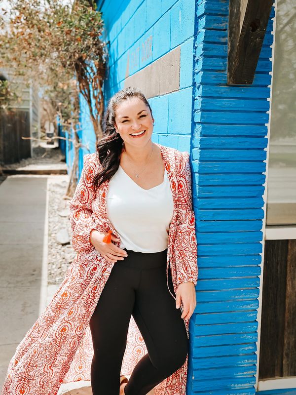 A smiling woman leans against a bright blue wall outdoors.