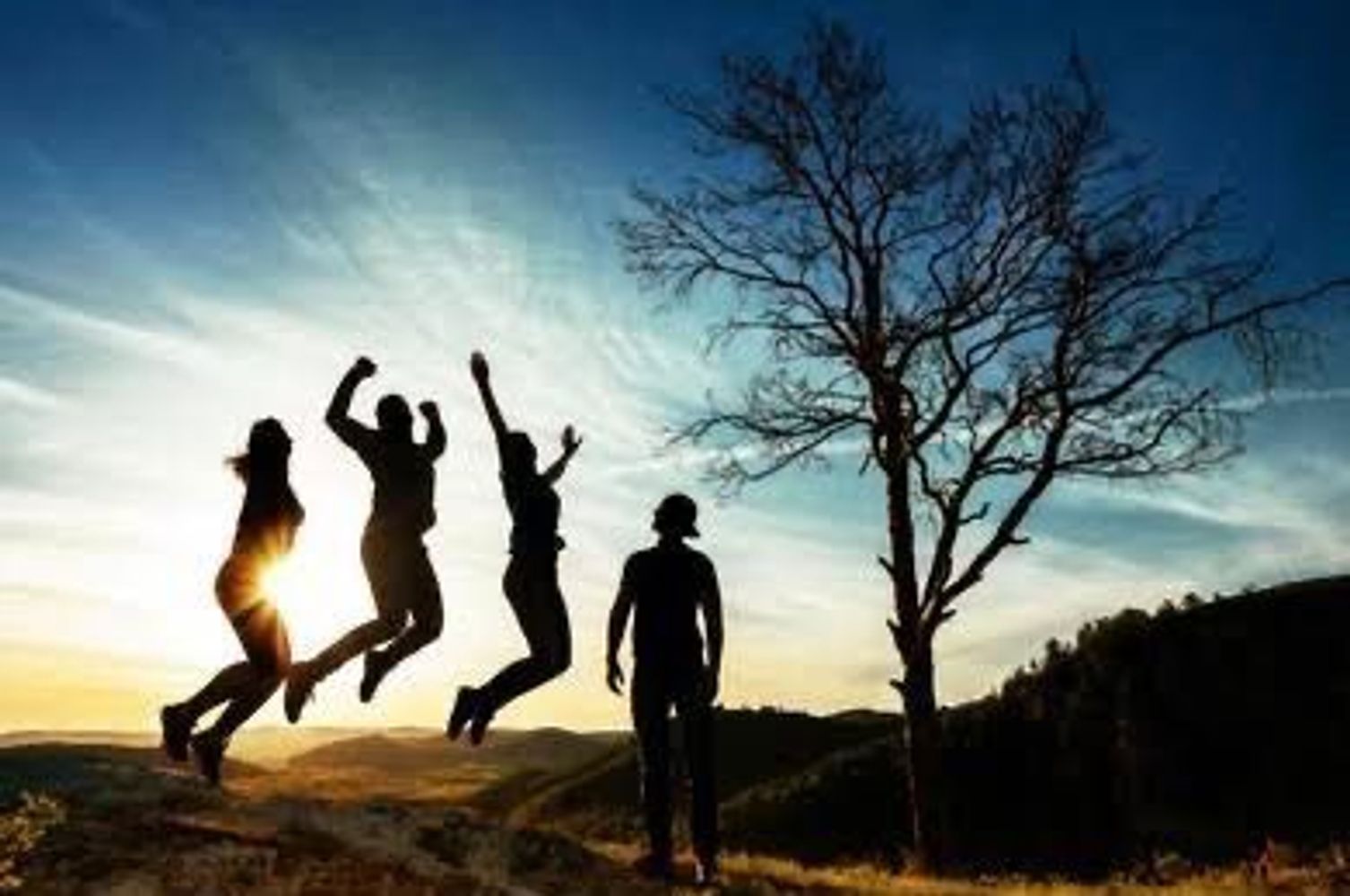 Four people enjoying sunset with three jumping and one standing by a tree.