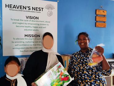 Three women smiling and holding items inside a child care center with a mission poster in the background.