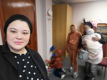 A woman takes a selfie with three children playing and standing in a room.