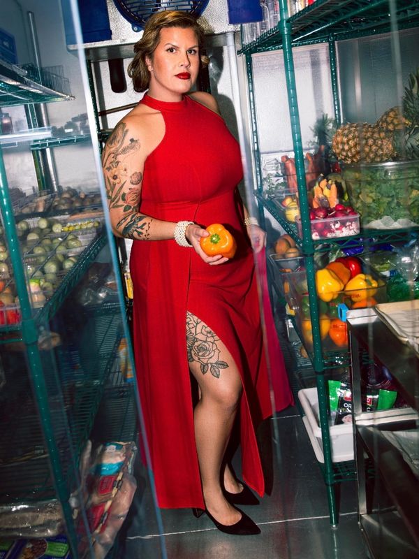 Woman in a red dress holding a bell pepper in a storage room full of fresh produce.