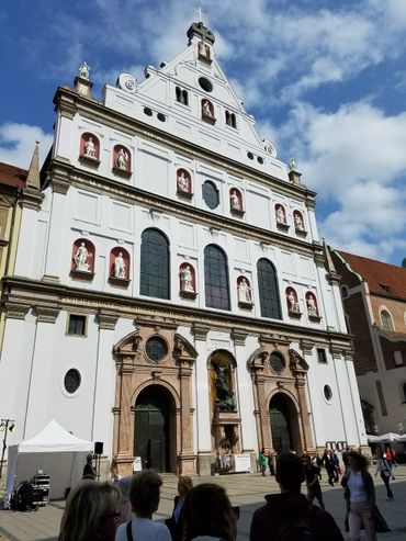 Facade of a historic white church with statues and large arched doors under a blue sky.