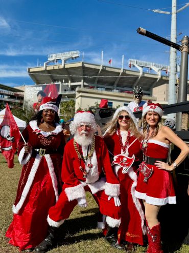 Four people dressed in festive red and white Tampa Bay Buccaneers holiday outfits outdoors.