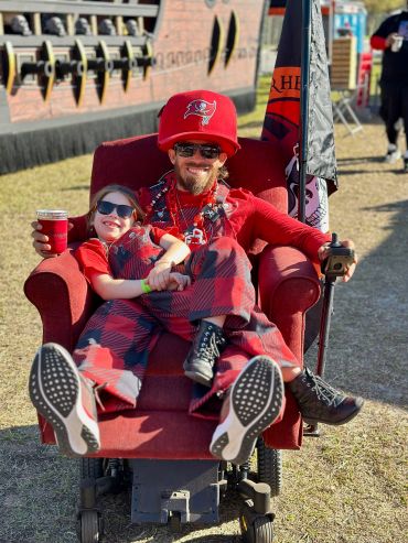 A man and child in Tampa Bay Buccaneers gear sitting on a red reclining chair outdoors.