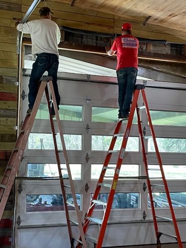 Two men working on a garage door while standing on ladders.