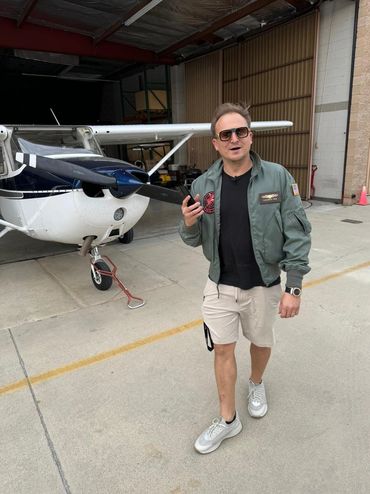 Man in aviator jacket and sunglasses walking near a small airplane.