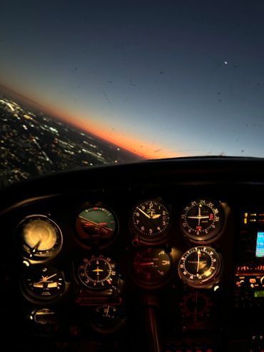 View from an aircraft cockpit at sunset with illuminated instrument panel.