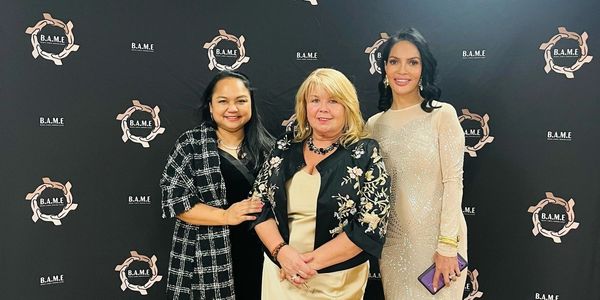 Three women posing in elegant dresses in front of a B.A.M.E backdrop at an event.