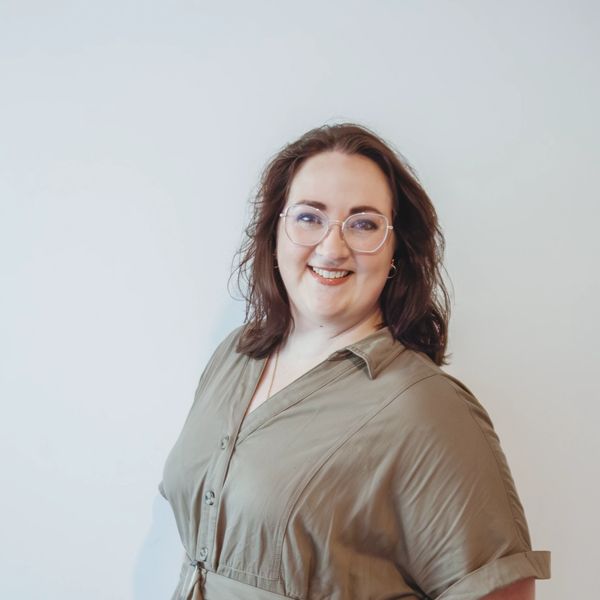 Smiling woman with glasses in a casual olive-green shirt against a plain background.