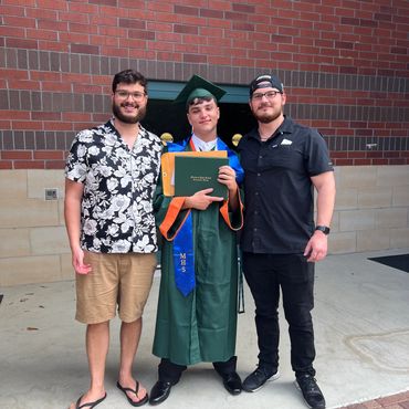 Three young men posing together, one in a graduation gown holding a diploma.