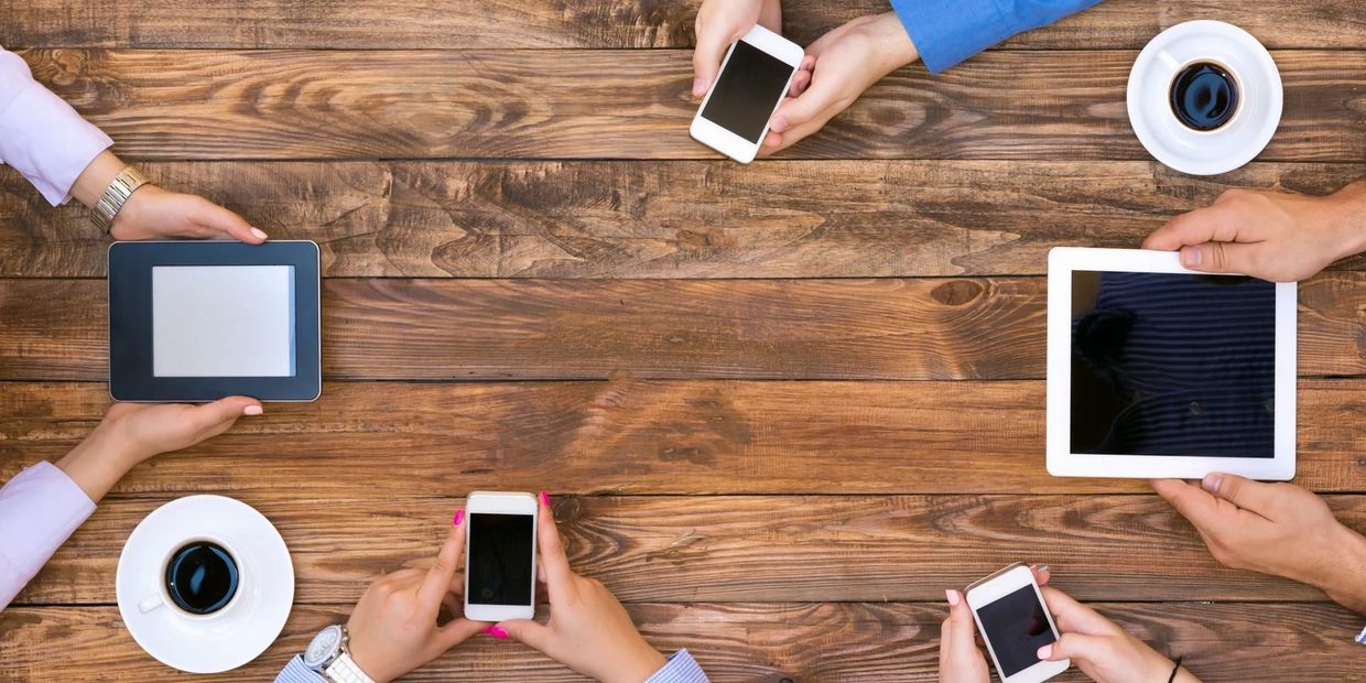 People holding smartphones and tablets around a wooden table with coffee cups.