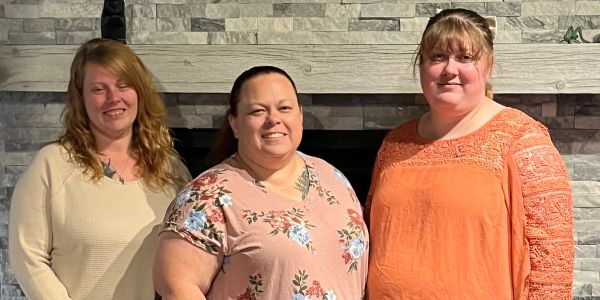 Three women smiling in front of a stone fireplace.