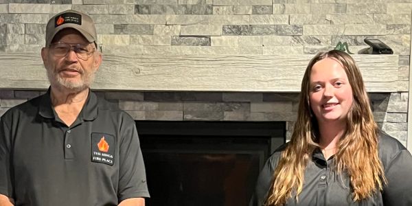 Two people in black shirts standing indoors by a fireplace.