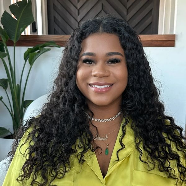 Smiling woman with curly hair wearing a yellow shirt and layered necklaces.