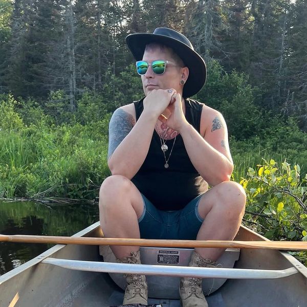 Person in sunglasses and hat sitting in a canoe on a calm water body surrounded by greenery.