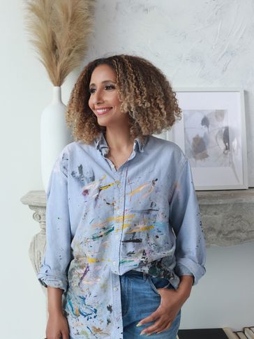 A smiling woman in a paint-splattered shirt stands casually indoors.