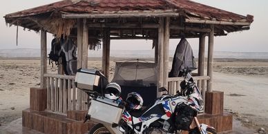 Motorcycle parked in front of a rustic hut on a sandy beach.