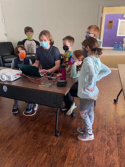 Staff member and 6 kids, all wearing face masks, around a computer and projector.