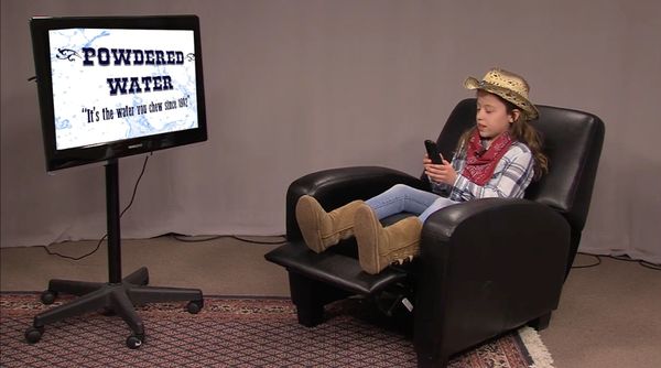 Cowgirl sitting in a recliner with phone in hand and "powdered water" on the TV screen