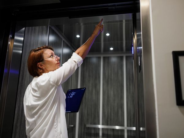 A person wearing a white shirt points upwards while holding a tablet in a modern, well-lit elevator.