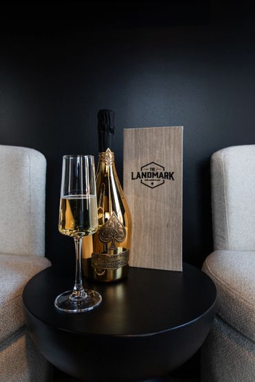 A glass and bottle of champagne on a black table between two beige chairs.