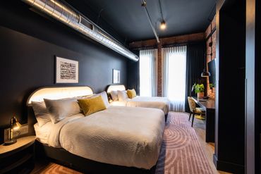 Modern hotel room with two beds, dark walls, and bright natural light from large windows.