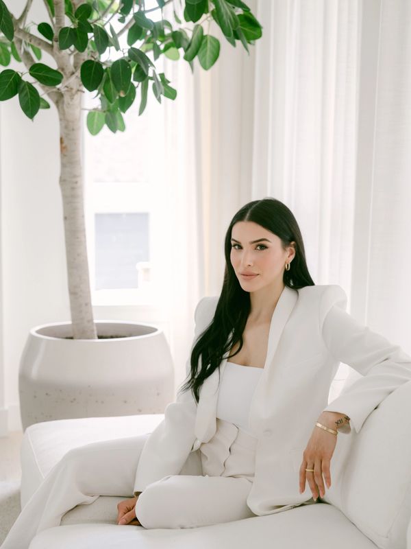 A woman in a white suit lounges on a white couch in a bright room with a large potted plant.