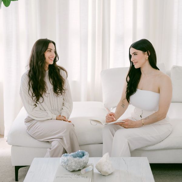 Two women having a relaxed conversation on a white couch in a bright room.