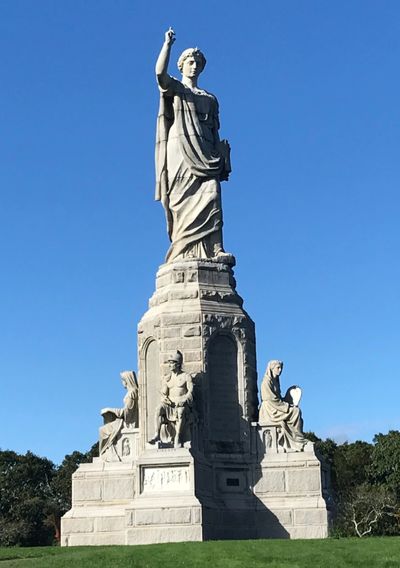 A tall stone statue stands against a clear blue sky on a grassy hill.