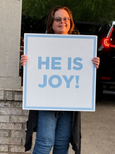 Woman holding a sign that says 'HE IS JOY!' with a smile.