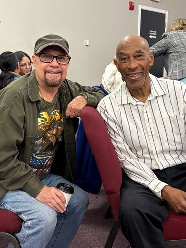Two men smiling, sitting on red chairs in a meeting room.