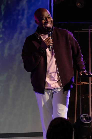 A man joyfully speaks into a microphone on stage with colorful lighting.