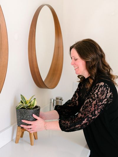 Female home organizer, in black, placing a plant between two mirrors on a bathroom vanity.