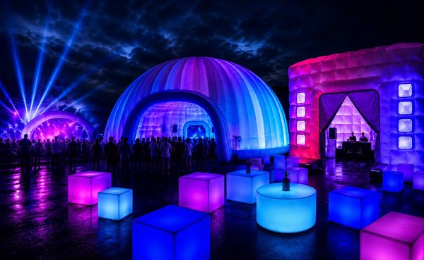 Colorful illuminated outdoor event with crowds and glowing furniture.
