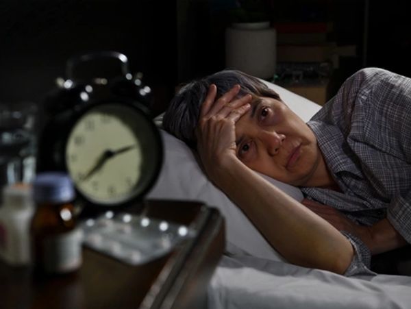 Elderly woman lying in bed wide awake at 2:30 AM. Bedside table holds a clock and sleeping pills