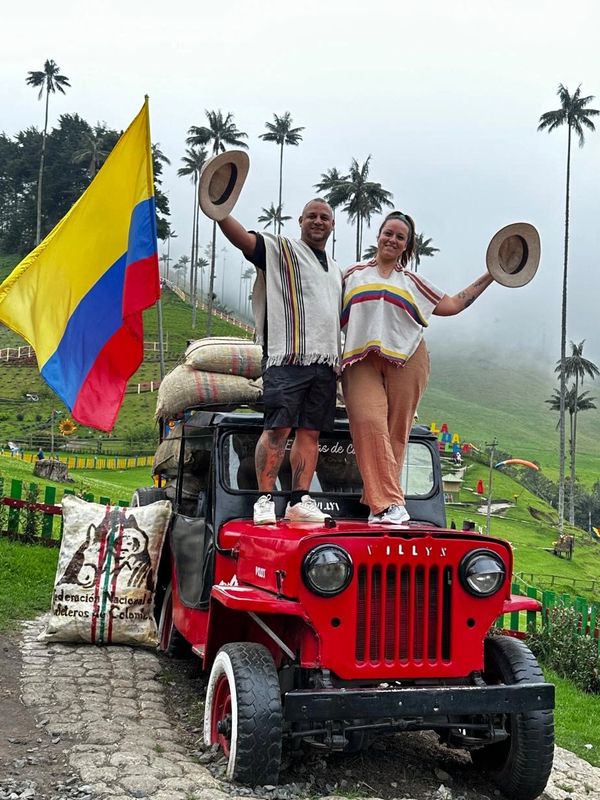 A joyful couple stands on a red Willys jeep with Colombian flag and coffee sacks in a green mountainous landscape.