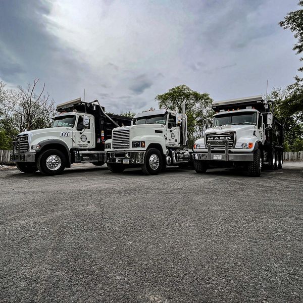Three white Mack dump trucks parked side by side on an asphalt lot under a cloudy sky.